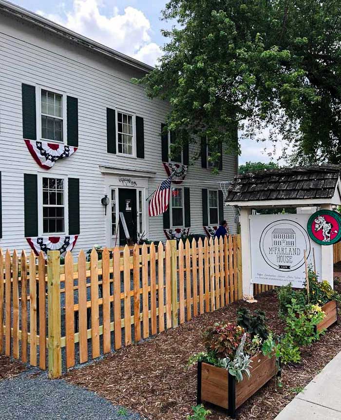 An outdoor sign displaying the McFarland House Logo with a white building to the left of the sign and wooden picket fence.