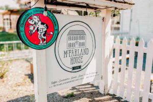 An outdoor sign displaying the McFarland House Logo with a white picket fence behind the sign.