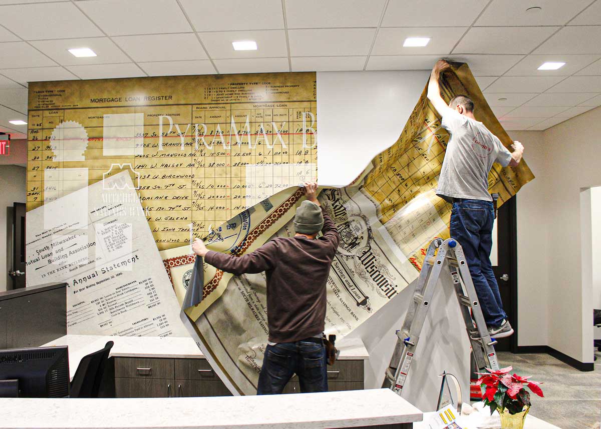 Two men installing PyraMax Bank's custom adhesive wall fabric behind a rounded front desk.