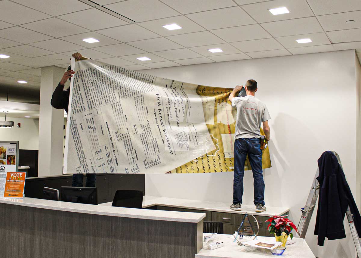 Two men installing PyraMax Bank's custom adhesive wall fabric behind a rounded front desk.