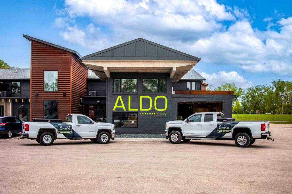 Two pickup trucks with custom graphics facing one another in front of a building displaying "ALDO".