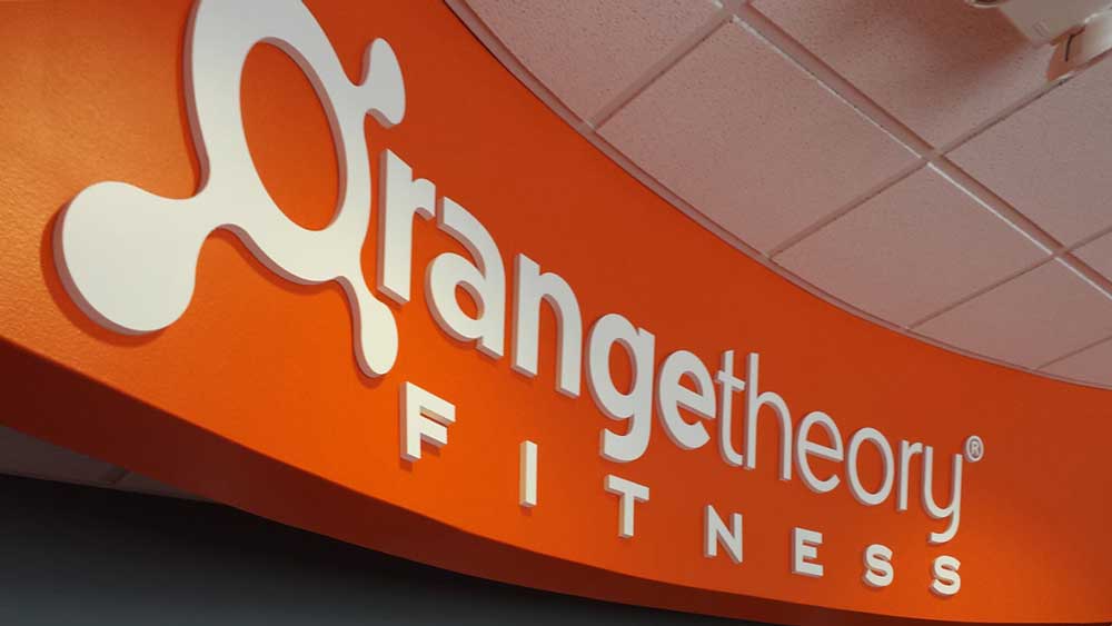 Orange Theory Signage featuring 3D white channel letters on a curved bright orange wall inside an Orangetheory Fitness location, viewed from below.