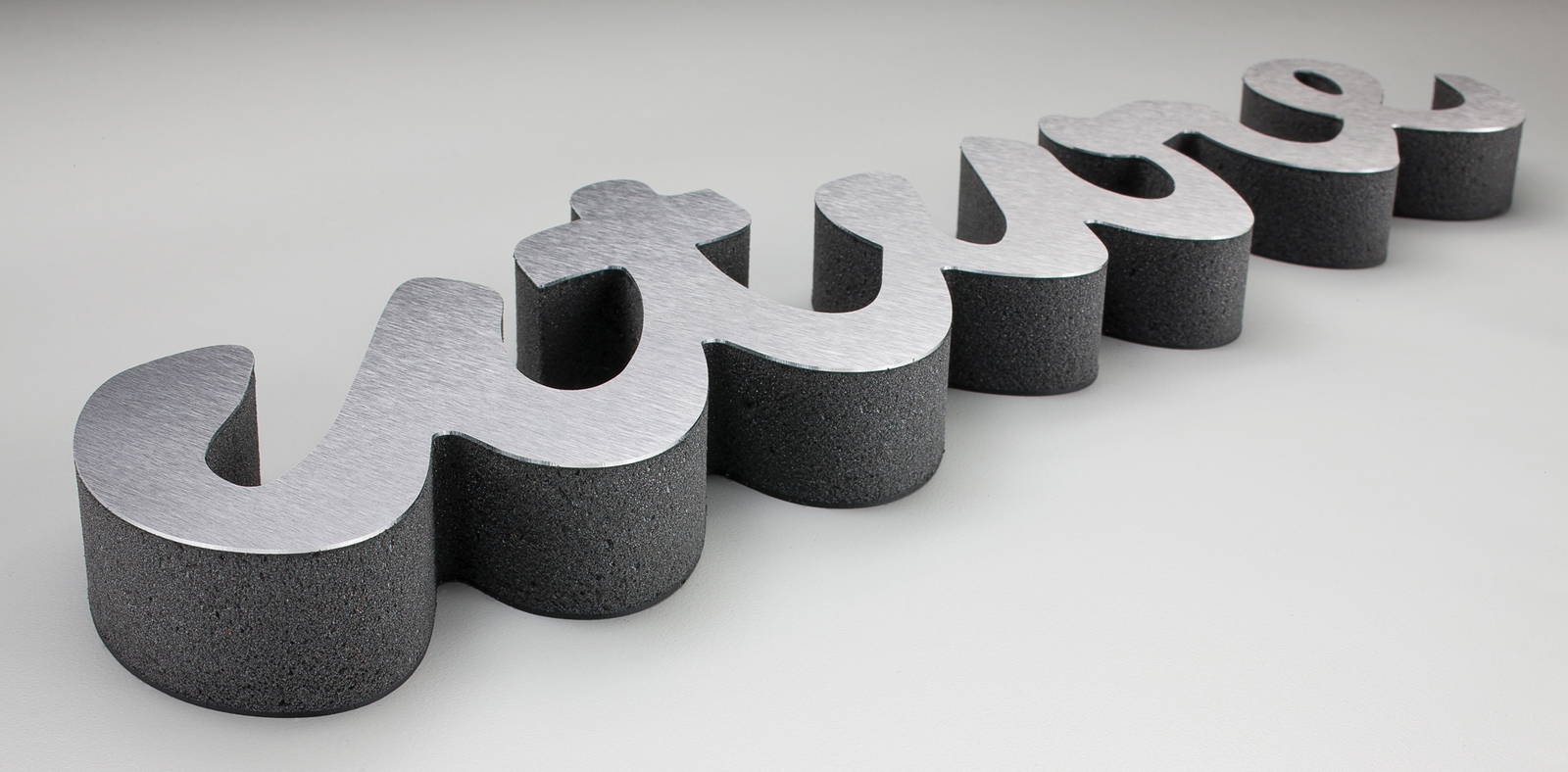 Laser-cut dimensional signage spelling the word “future” in a cursive script, featuring a brushed metal face and thick textured black sides, displayed on a light gray surface.