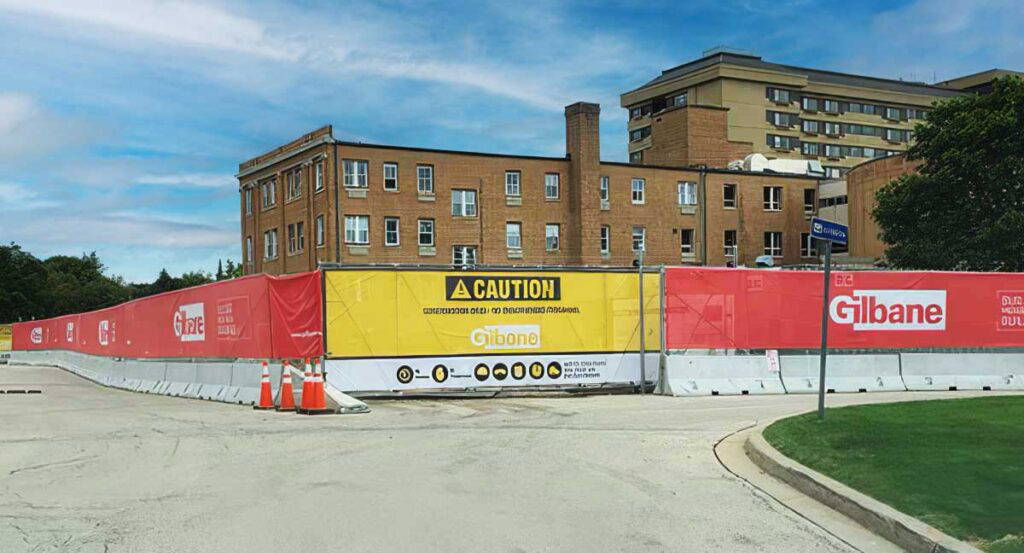 Red and yellow hanging construction banners on a chain-link fence in front of a site with a large brick building.