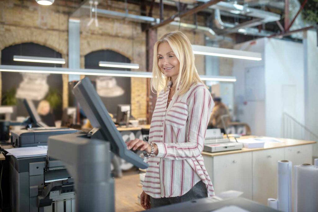 Woman with blonde hair in a long sleeve shirt using an industrial printer.