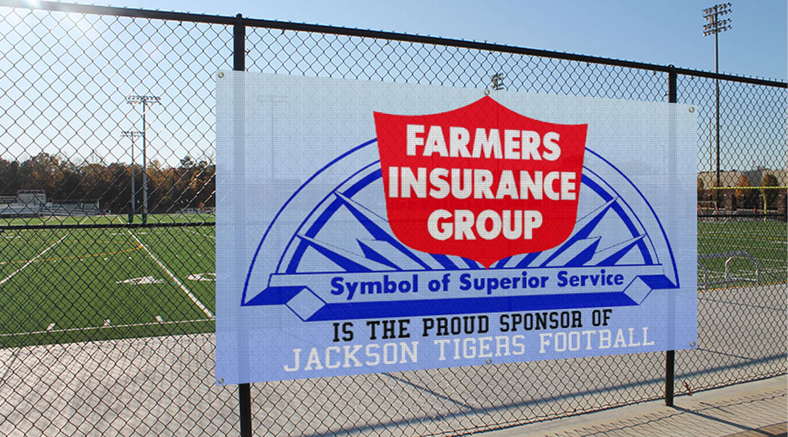Mesh banner for Farmers Insurance Group attached to a chain-link fence at a football field, displaying the company logo and the message “Symbol of Superior Service” and “Proud Sponsor of Jackson Tigers Football.”