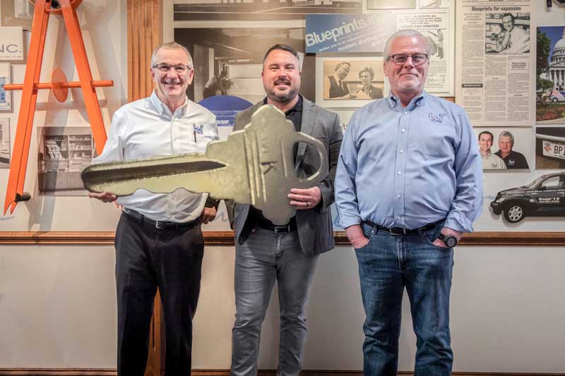 3 men standing in front of BPI Color's history wall holding a large, gold key.