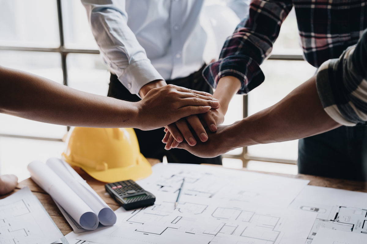 4 hands stacked above a white table with a hardhat and a rolled up blueprint,