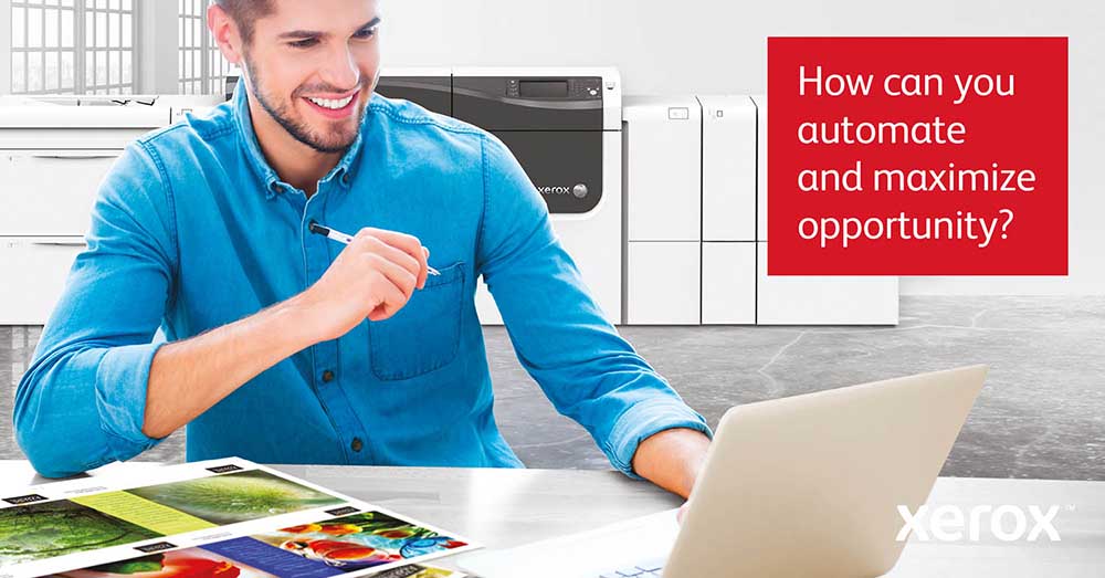 HP® DesignJet® XT950 MFP 11 Xerox Print Automation – Smiling professional at a desk with a laptop and vibrant print samples, with a Xerox production printer in the background.