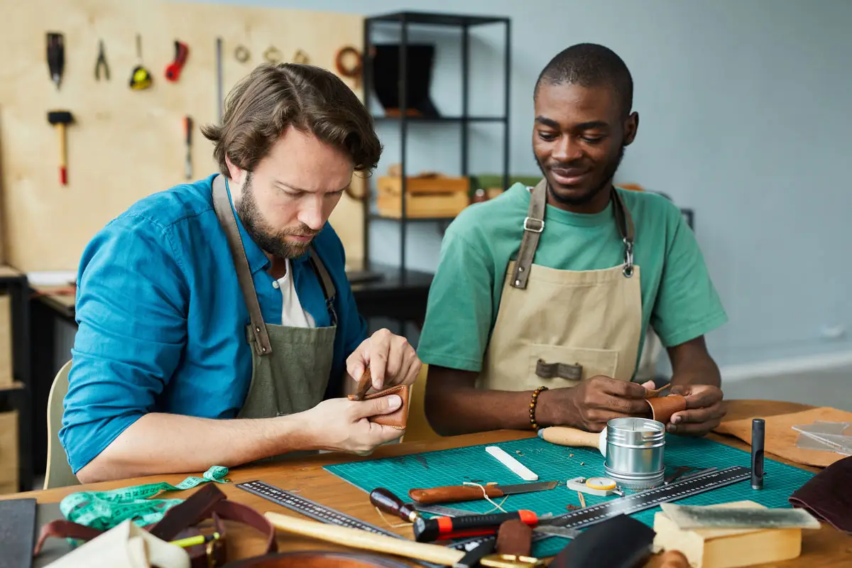 BPI Color's 2024 Year in Review 14 Woodworking Studio scene of two men in aprons crafting leather pieces by hand, surrounded by tools, rulers, and materials on a shared workshop table.