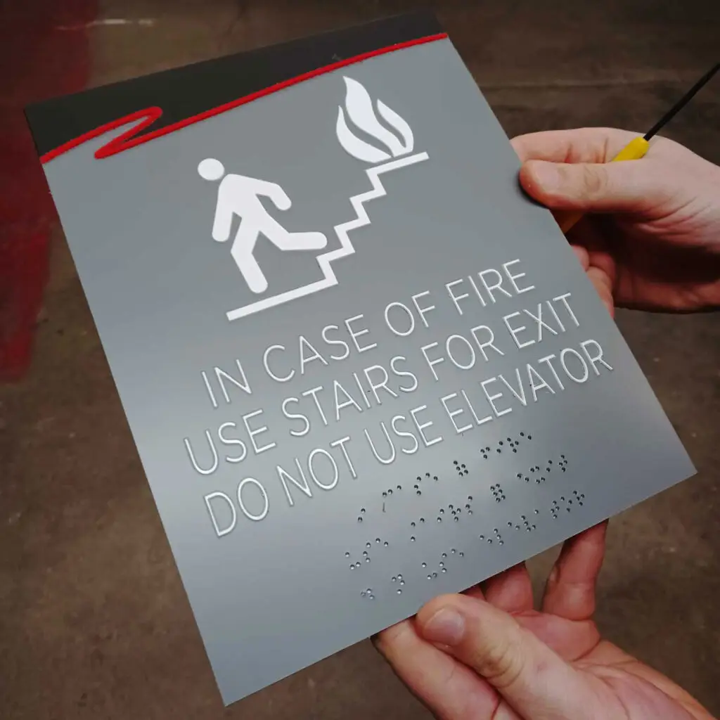 ADA and Wayfinding Signage showing a tactile and braille fire safety sign that reads "In Case of Fire Use Stairs for Exit Do Not Use Elevator" being held by a person.