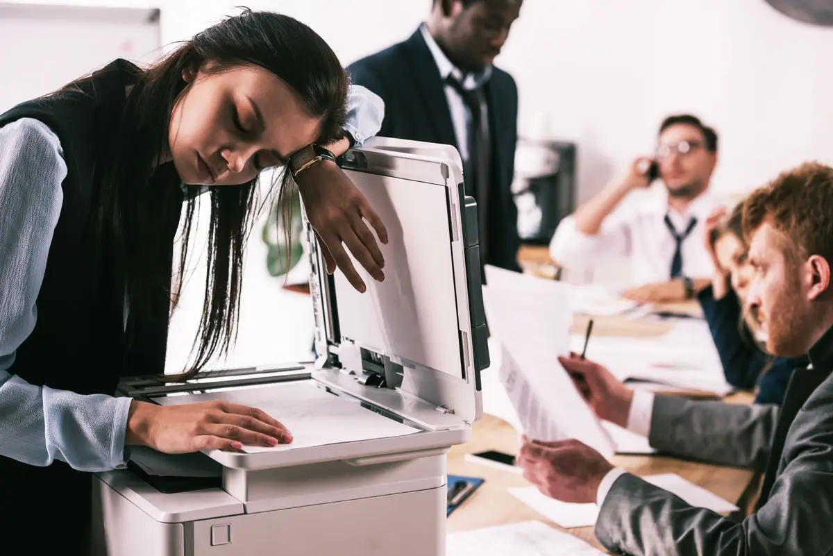 Bricked Printer scene showing a frustrated office worker resting her head on a broken printer, while stressed colleagues deal with paperwork and calls in the background.
