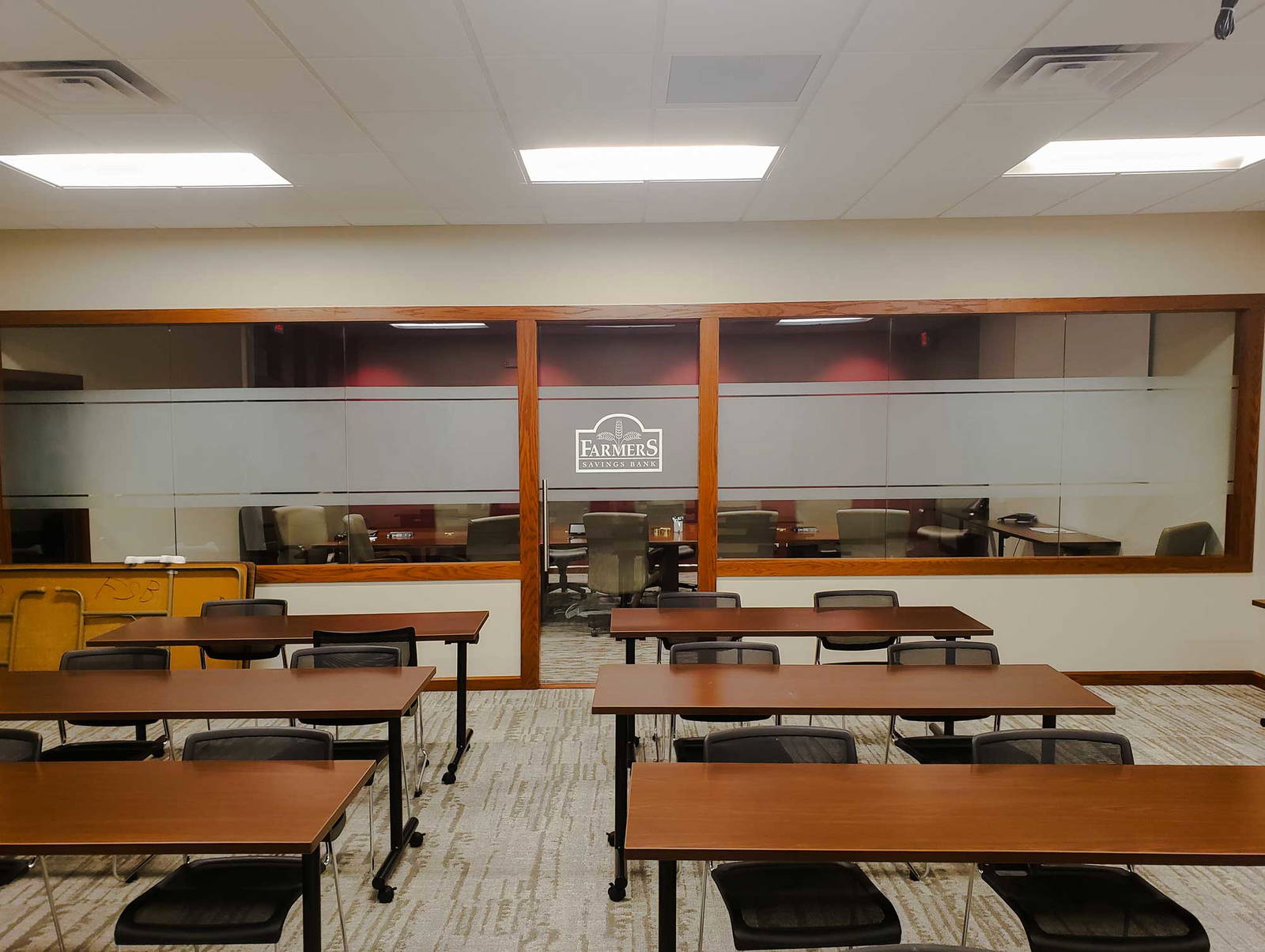 Training or classroom area with long tables and chairs facing glass-walled offices labeled with the Farmers Savings Bank logo, providing a view into an adjacent conference room.