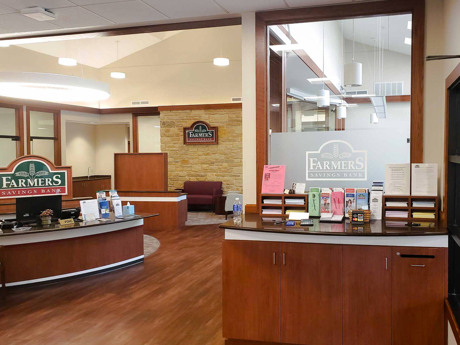 Interior view of Farmers Savings Bank, featuring a warm, wood-accented reception area with informational brochures, branded signage, a frosted glass window, and a customer service desk.