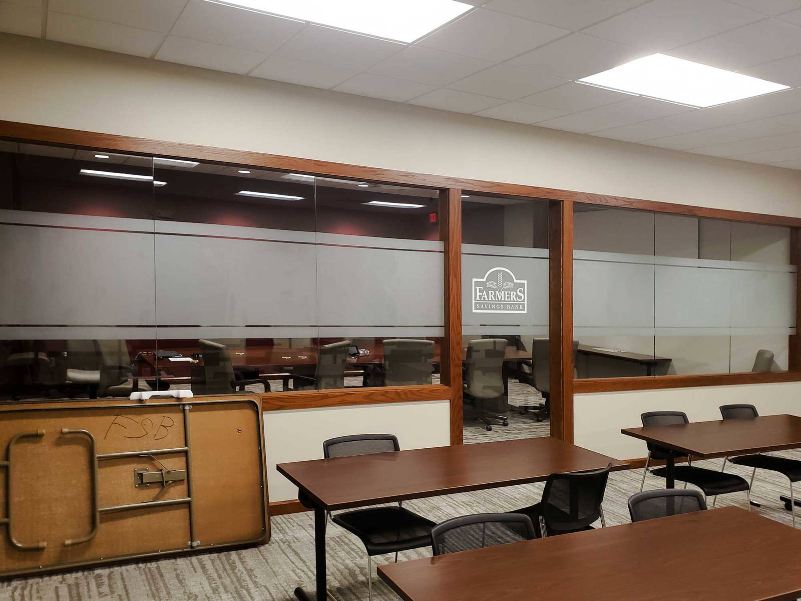 Conference-style meeting room with glass walls branded with the Farmers Savings Bank logo, viewed from a classroom-style space with rows of tables and chairs.