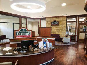 Welcoming interior of Farmers Savings Bank featuring a curved reception desk, branded signage, brochure displays, stone accent wall, and waiting area with armchairs.