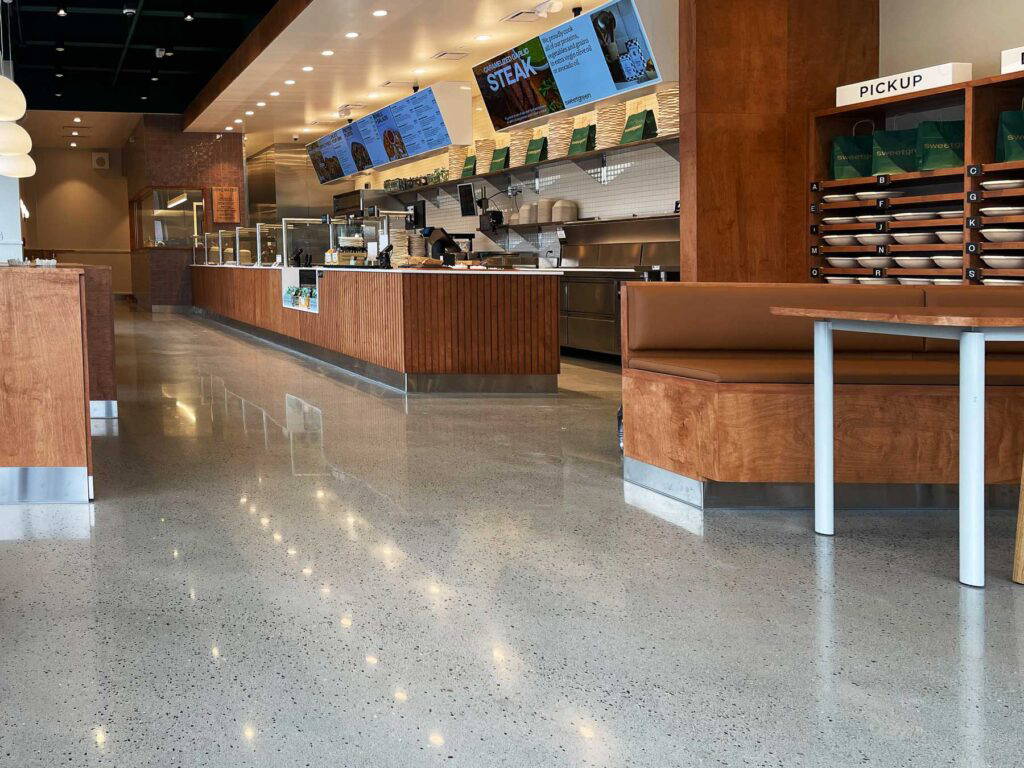 Modern restaurant interior featuring polished concrete floors, wood-accented counters and seating, digital menu displays, and a designated order pickup area.