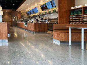 Modern restaurant interior featuring polished concrete floors, wood-accented counters and seating, digital menu displays, and a designated order pickup area.