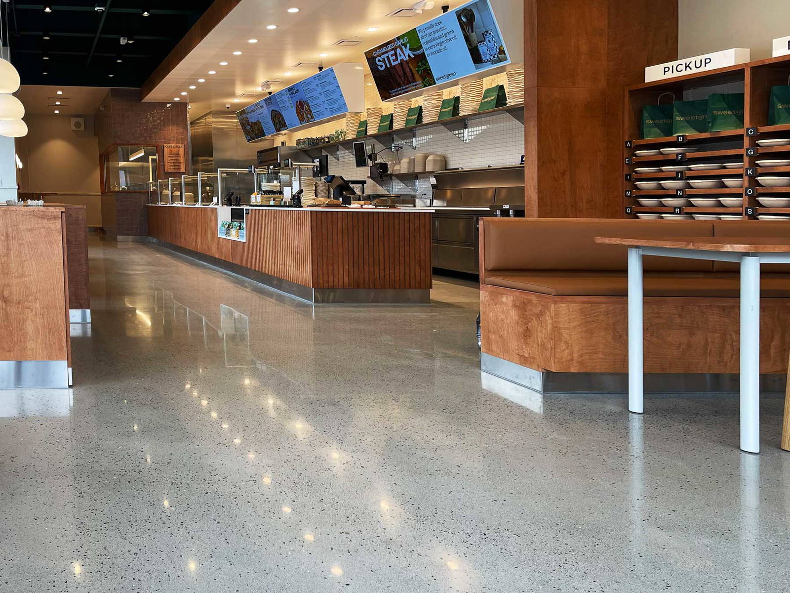 Modern restaurant interior featuring polished concrete floors, wood-accented counters and seating, digital menu displays, and a designated order pickup area.