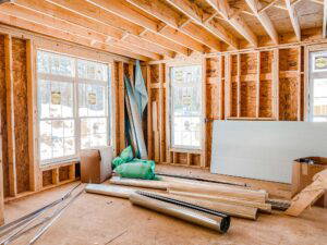 Interior view of a residential construction site showing exposed wooden framing, new window installations, insulation materials, and construction supplies on the floor.
