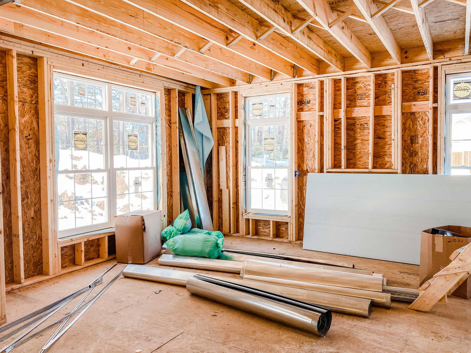 Interior view of a residential construction site showing exposed wooden framing, new window installations, insulation materials, and construction supplies on the floor.