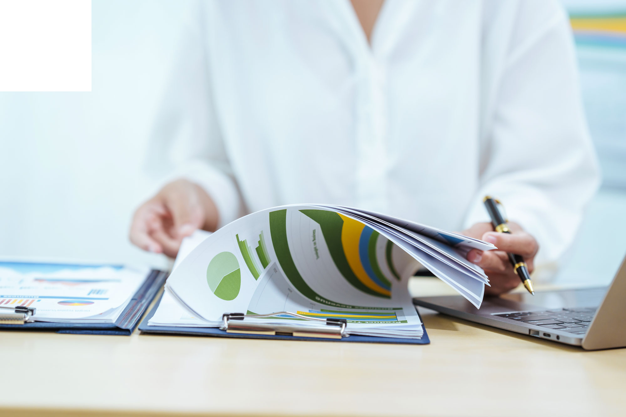 Business Printing materials including colorful charts and reports in a binder, reviewed by a person in a white blouse using a laptop and pen.