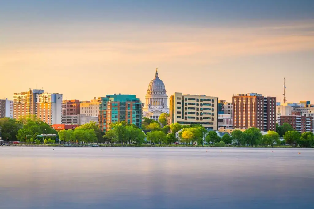 Madison Wisconsin skyline at sunset with the Wisconsin State Capitol building centered among modern buildings, viewed across a calm lake with green trees in the foreground.