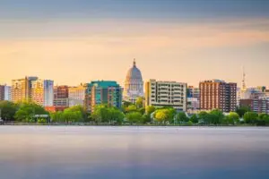 Madison Wisconsin skyline at sunset with the Wisconsin State Capitol building centered among modern buildings, viewed across a calm lake with green trees in the foreground.