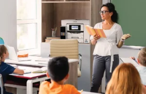 Middle ages female teacher standing with a book in her hands in front of the class with an Epson printer in the background.