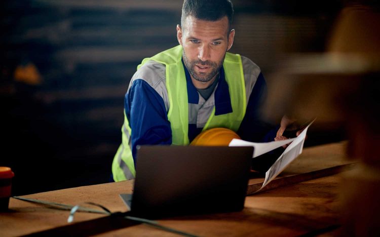 Close-Out Docs image showing a construction worker in a high-visibility vest reviewing project documents on a laptop at a worksite, symbolizing end-of-project reporting.