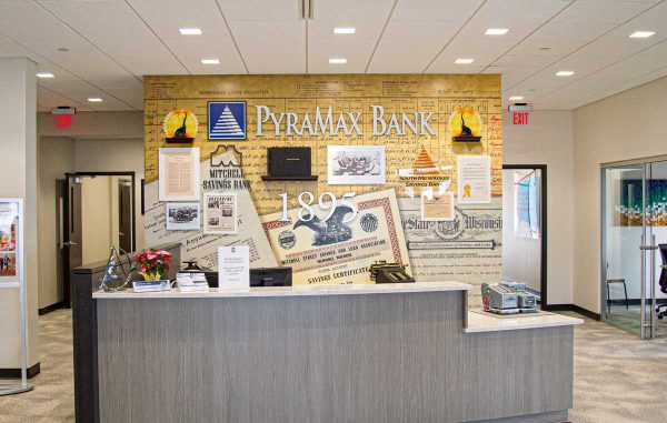 Environmental Graphics - PyraMax Bank History Wall featuring a visually layered collage of vintage banking documents, photos, and signage behind the reception desk.