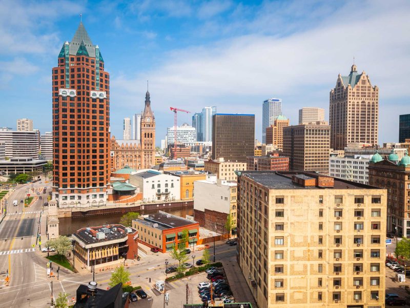 Milwaukee, WI downtown skyline on a bright summer day featuring a mix of historic and modern architecture, with prominent buildings like the Milwaukee City Hall and 100 East Wisconsin under a blue sky.