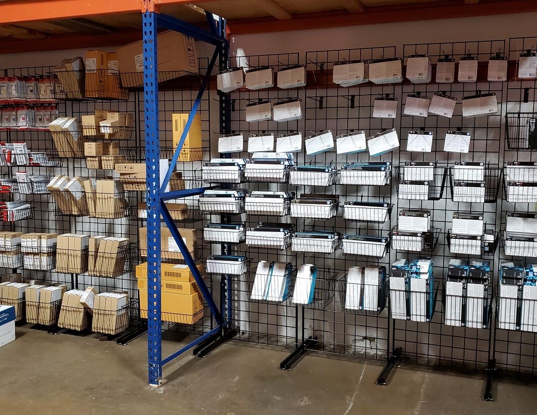 Printer Supplies Printer Supplies inventory wall featuring organized HP and Canon ink cartridges, toner boxes, and packaging materials displayed in wire baskets and shelving.