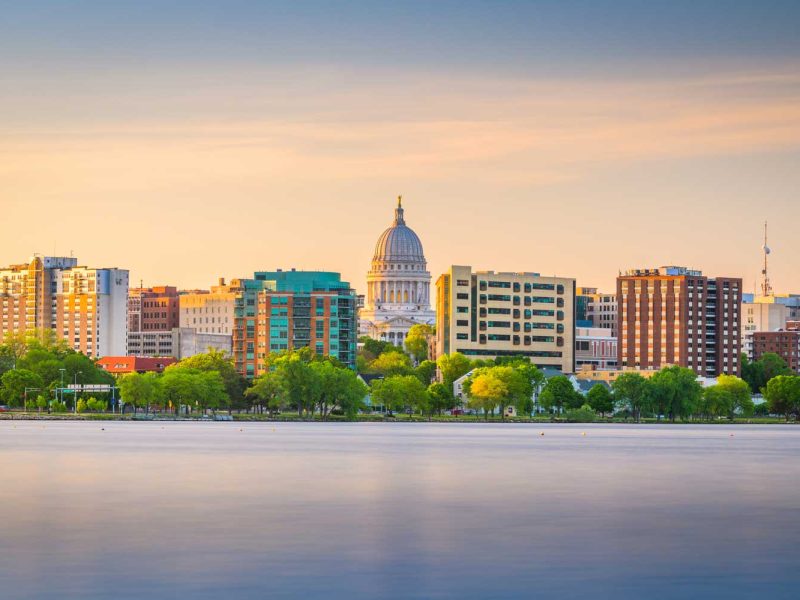 Madison Wisconsin skyline at sunset with the Wisconsin State Capitol building centered among modern buildings, viewed across a calm lake with green trees in the foreground.