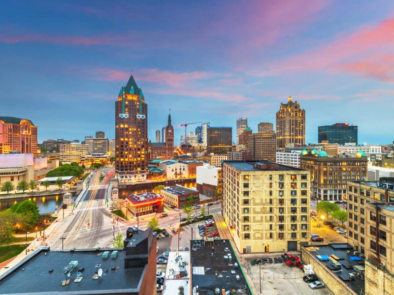 Milwaukee, WI skyline at dusk showcasing downtown buildings, bridges, and city lights beneath a vibrant pink and blue sky.
