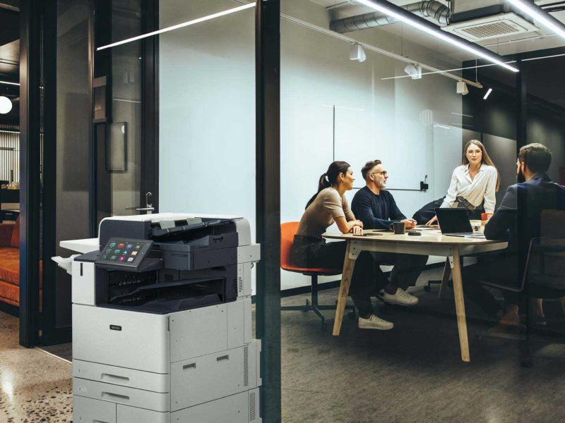 Xerox Lifestyle image featuring a modern office with a Xerox multifunction printer in the foreground and a team of professionals collaborating in a glass-walled meeting room.
